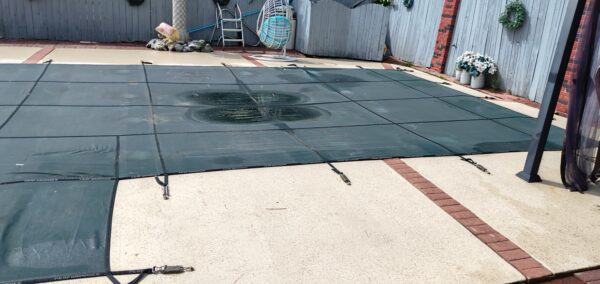 Backyard with a large black pool safety cover secured over a concrete patio. Straps and anchors visible along edges.