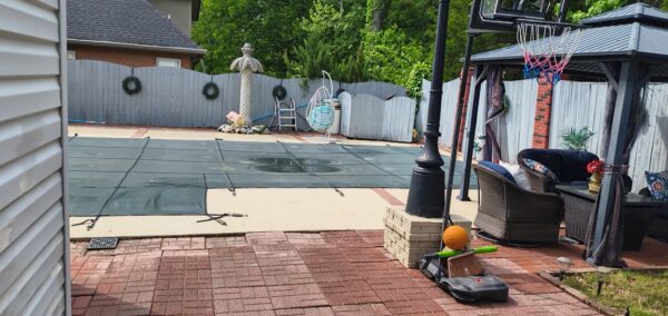 Backyard with a covered pool, brick patio, and wicker seating under a gazebo in a fenced yard with trees behind it.