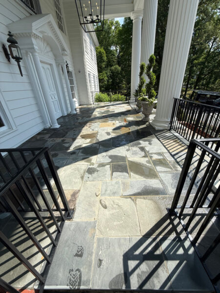 Stone slate porch with white columns, black railings, and potted topiary on a sunny veranda.