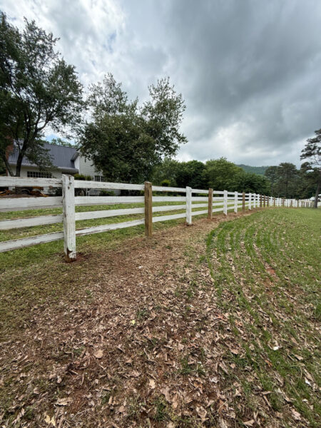 White split-rail fence along a curved grassy field with leaf-covered ground and a distant house under a cloudy sky