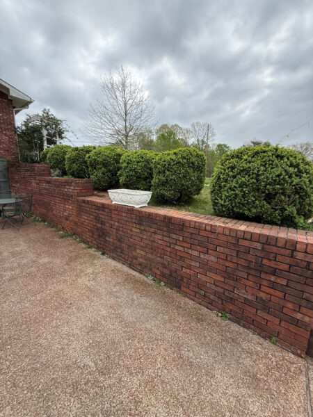 Brick wall with a white decorative planter on top, green hedges behind, cloudy sky above.