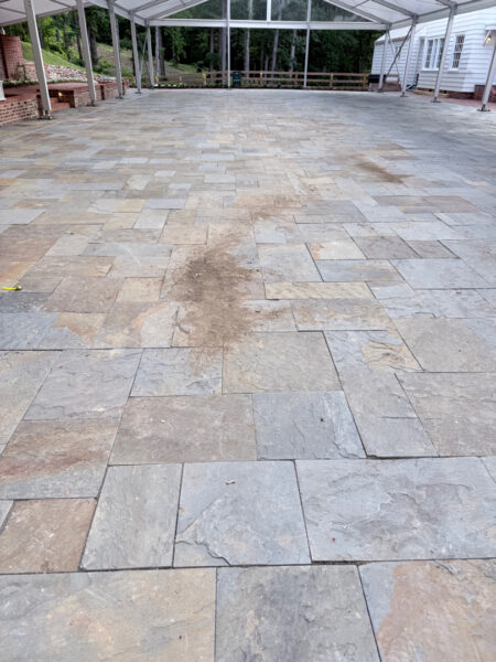 Large stone-paved outdoor patio or courtyard under a metal-framed canopy, with dirt patches and a white building to the side.