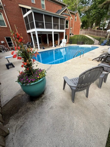 Backyard pool area with blue water, concrete deck, and Adirondack chairs near a brick house with a screened porch above the patio garden bed.