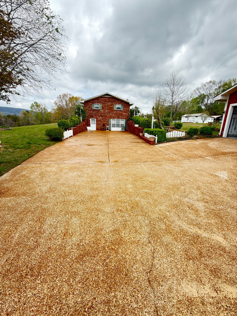 Brick two-story house at end of a wide gravel driveway, with white front door and trimmed greenery along brick walls under a cloudy sky.
