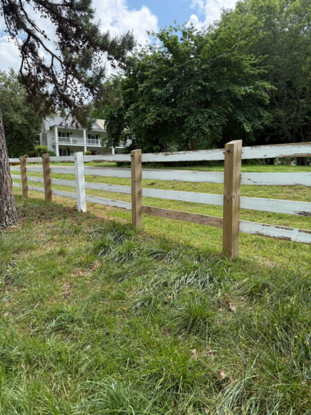 Wooden split-rail fence along a grassy yard with trees and a two-story house in the background under a blue sky.