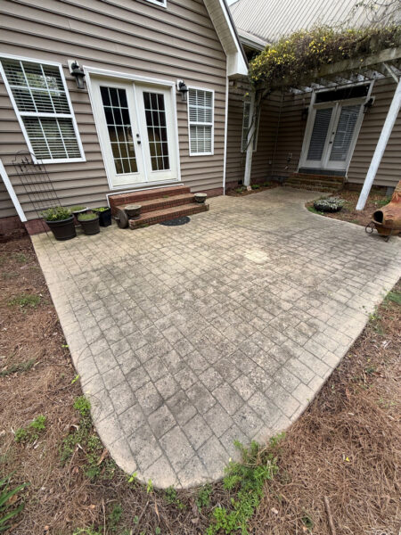 Paved backyard patio with interlocking concrete pavers, white French doors, and three brick steps leading to the door, with several plant pots along the wall. A clay chiminea sits on the right side of the space.