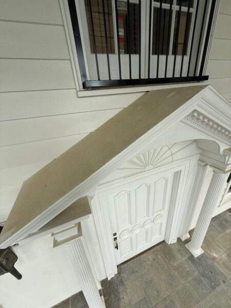 White exterior doorway with ornate pediment, fluted columns, and a slanted concrete landing above the door; window with black bars visible above.