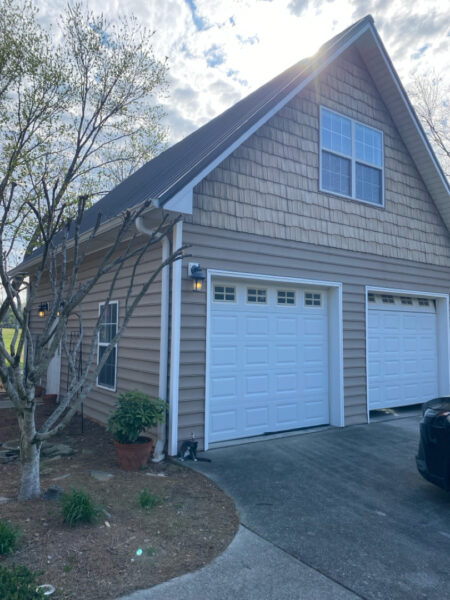 Two-car garage with a steep gable roof, beige siding, and a cat sitting by the driveway near a potted plant in a suburban yard.