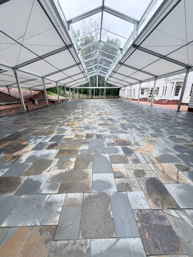 Long glass-roofed walkway with slate stone tiles, brick walls, and a white building on the right in the distance.