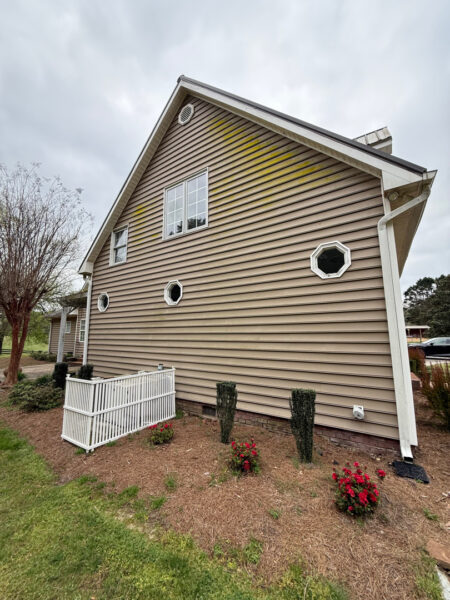Side view of a beige house with a steep gabled roof, four small octagonal windows, white trim, and a mulched yard with red roses and trimmed shrubs.