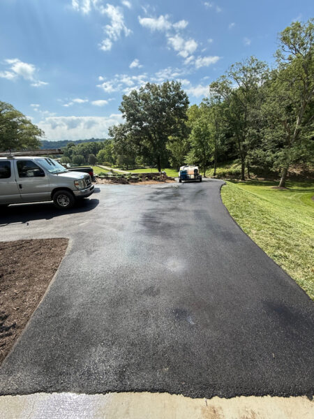 Freshly paved black asphalt driveway curves through a green yard with a white van parked on the left and a small trailer in the distance.
