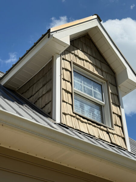 Dormer window on a beige wooden-shingle house under a blue sky with white trim