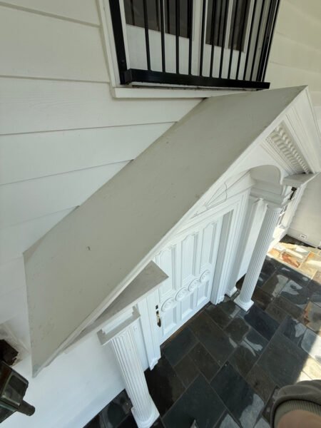 White front porch entry with a slanted triangular landing, ornate fluted columns, and a white paneled door. A black railing sits above a window.