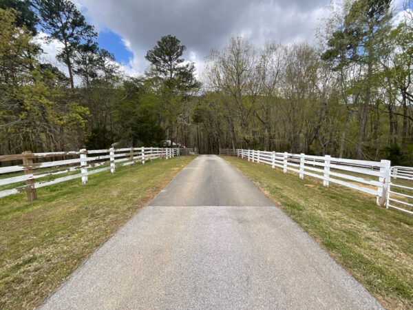Paved lane flanked by white split-rail fences, grassy shoulders, and leafless trees under a cloudy sky.