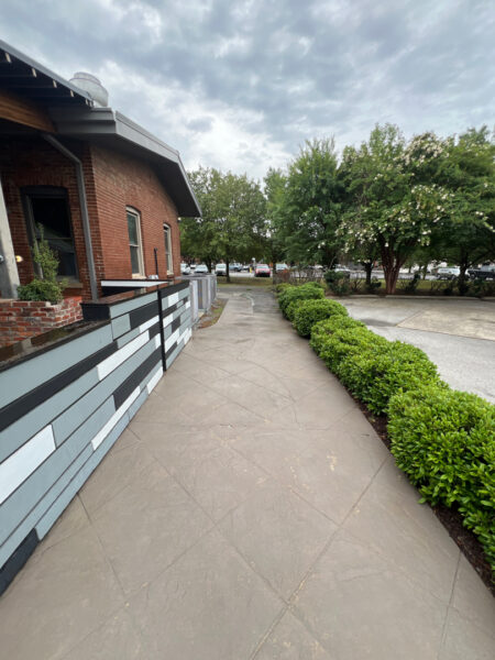 Sidewalk beside a brick building with a striped decorative wall, green hedges on the right, and trees in the distance under a cloudy sky