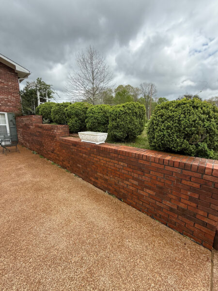 Brick red wall runs along a gravel patio, with a white decorative planter on top and dense green hedges beyond under a cloudy sky.