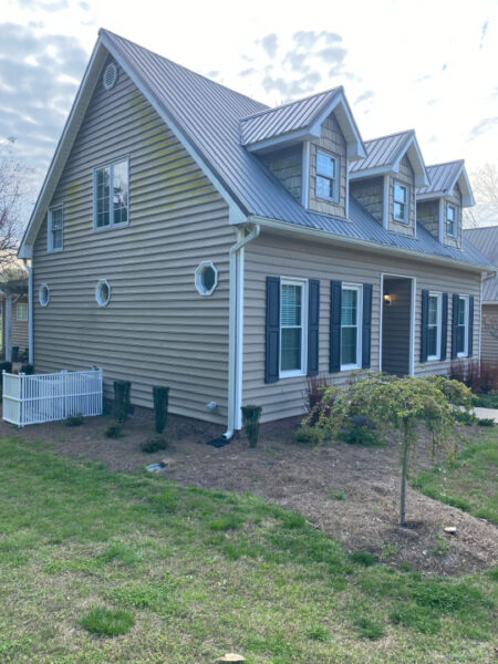 Two-story beige house with a metal pitched roof, blue shutters, dormer windows, and a small front porch, surrounded by shrubs and a grassy yard.