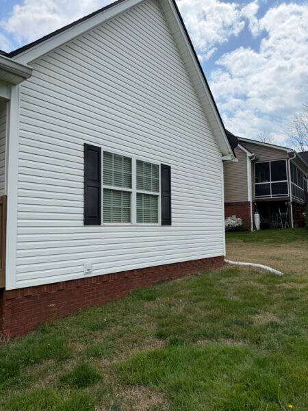 White house with vinyl siding, a double-hung window with black shutters, and a brick foundation curb; green lawn in foreground.