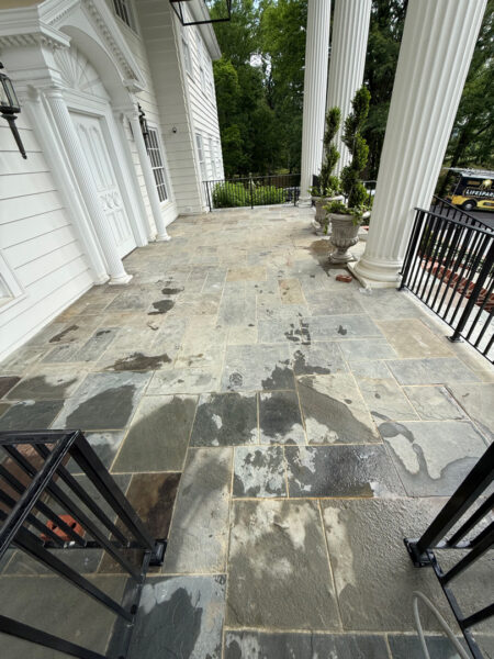Front porch with tall white columns and ornate trim, damp slate tiles, and a black railing overlooking greenery.