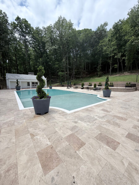 Outdoor pool area with beige tiled patio, a rectangular pool, and tall potted topiary plants; forested backdrop and small pool house to the left.