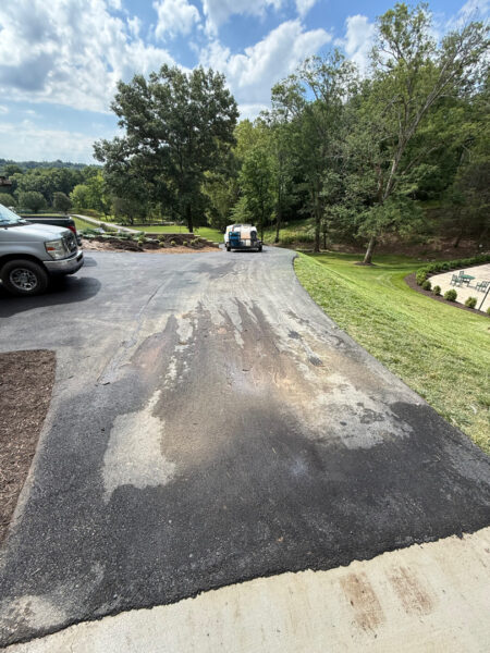 Paved driveway with worn, patchy asphalt stretching toward a small trailer in the distance, while a pickup sits on the left and trees line the background.