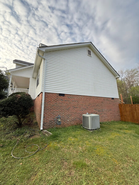 Rear corner of a white house with brick foundation, an outdoor AC unit, downspout, and a coiled hose on the lawn.