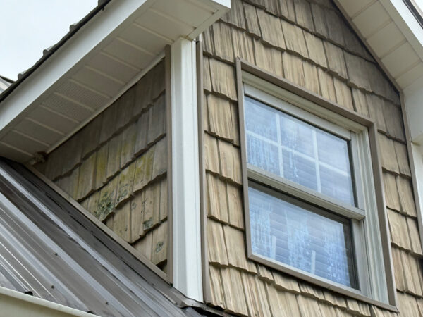 Wood-clad exterior with weathered wooden shingles surrounding a double-hung window under a sloped roof. The window has a white frame and is set in a brownish, aged siding section on a house corner.