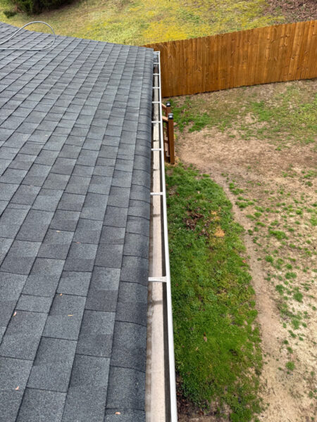 A rooftop seen from above with gray shingles, along the edge is a white gutter and fascia board, overlooking a yard and wooden fence in the background.