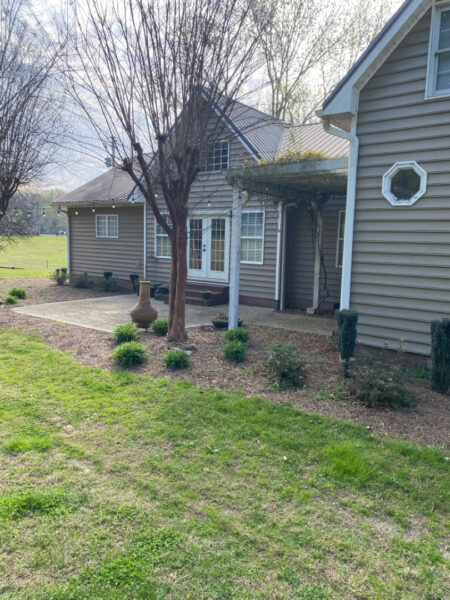 Exterior view of a beige siding house with a covered porch, a large tree in front, and a mulched garden bed with small shrubs.