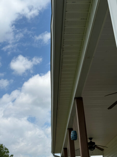 Side view of a white porch soffit and gutter with brown posts, a blue lantern hanging, and a ceiling fan against a blue sky with clouds.
