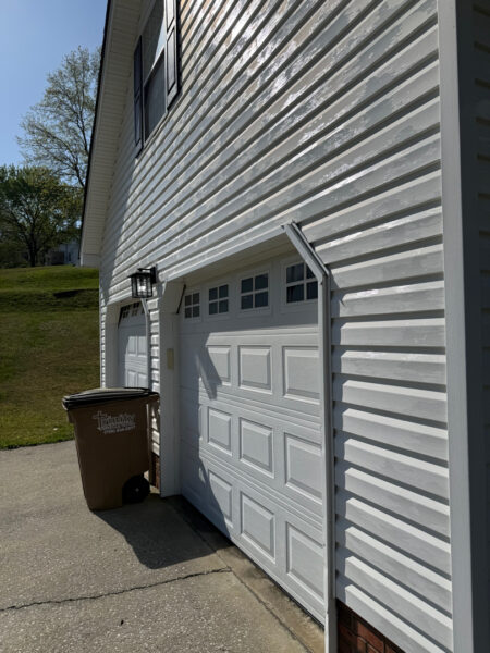 White vinyl-siding garage exterior with a white raised-panel door, a black lantern light, and a brown trash bin by the entrance in the driveway.