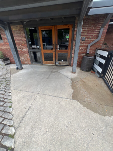 Wooden-framed glass double doors at a brick building entrance under a metal awning, with a wet concrete path.
