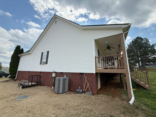 White house with brick foundation and a raised wooden deck on the right, stairs leading down, with lawn tools and an AC unit nearby in the yard.