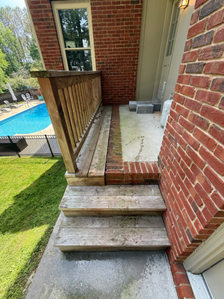 Wooden steps and a small porch lead up to a green door on a brick house; in the background is a fenced backyard with a blue pool and patio furniture.