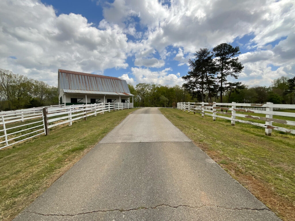 Paved country road flanked by white wooden fences, a white barn on the left, green pastures, and tall trees under a partly cloudy sky.