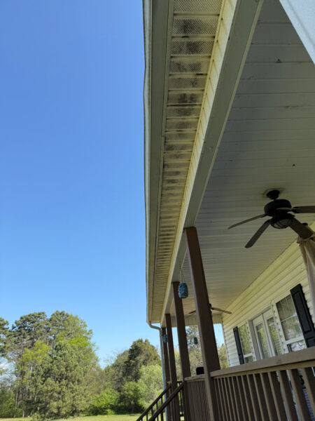 Covered porch with brown posts, a ceiling fan, hanging lanterns, and a blue sky with trees in the background.