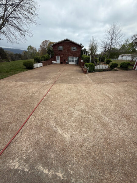 Brick two-story house at end of a wide concrete driveway with a red hose running along it; manicured yard and trees on either side under a cloudy sky.