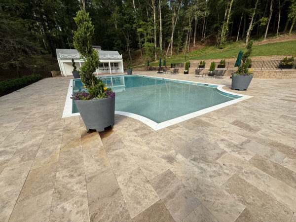 Rectangular outdoor pool surrounded by beige stone tiles, with large planters and a small white pool house in the background.