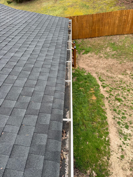 Aerial view of a gray shingled roof edge with a narrow gutter along the eave and a white downspout at the gutter line, overlooking a gravel yard and wooden fence in the background.