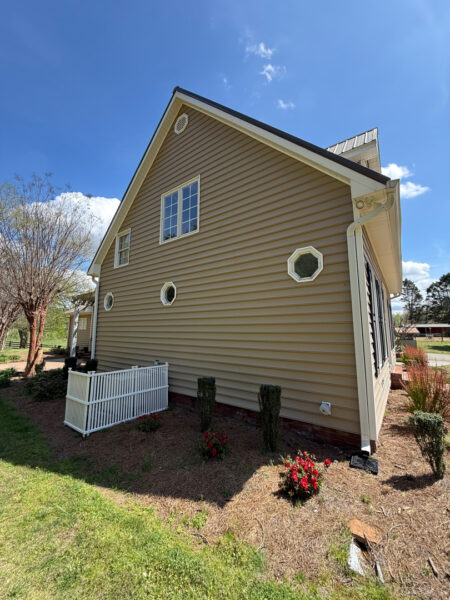 Beige, two-story house with white trim, octagonal windows, and a small white fence beside a mulched garden bed under a blue sky.