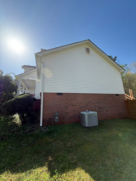Side view of a white house with brick foundation, outdoor AC unit on the lawn, and a coiled hose nearby under a bright sun.