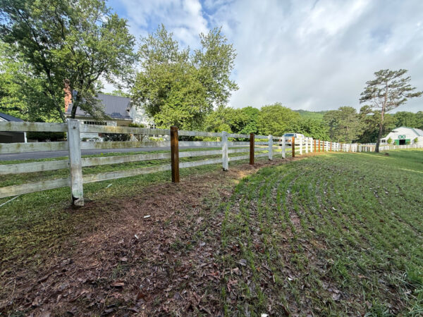 White split-rail fence along a grassy field with trees and a house in the distance under a partly cloudy sky.