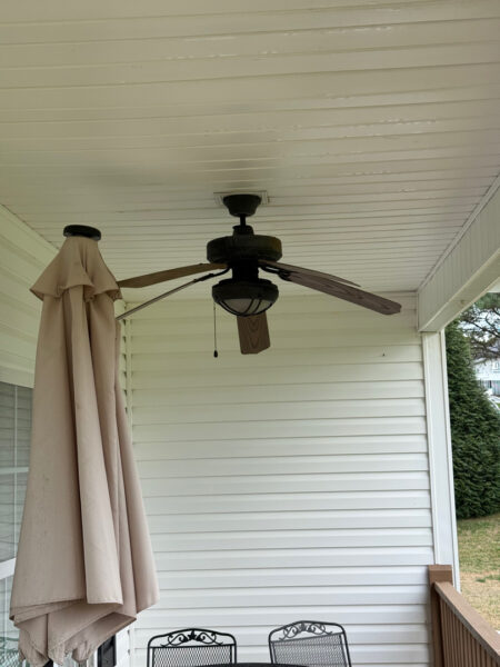 Ceiling fan with four blades above a white covered porch, beige patio umbrella folded to the side, and metal chairs along the wall.