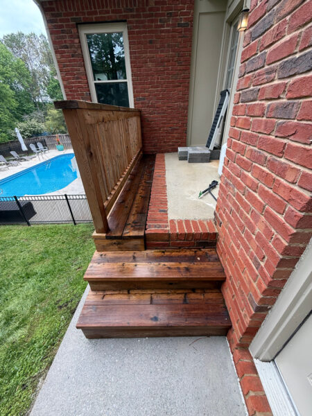 Wooden stairs and railing lead up to a brick house, with a concrete landing and pool area visible in the yard beyond the fence.