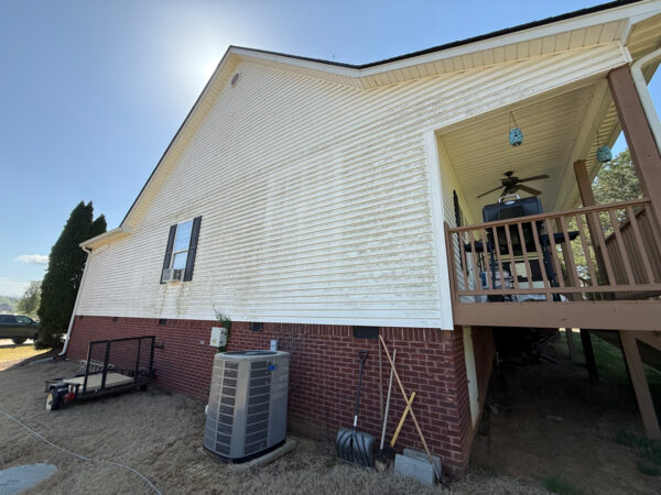 Side view of a white house with brick foundation, an outdoor air conditioner, and a raised porch with a grill and hanging lanterns on a sunny day.