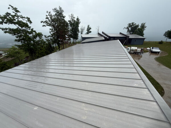 Overhead view of a long silver metal roof on a hillside, wet from rain, with trees and a small building complex in the distance and a curved driveway with vehicles nearby.