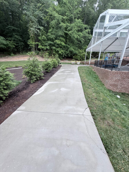 Concrete sidewalk curves through a landscaped area toward a glass-walled pavilion, with trees in the background.