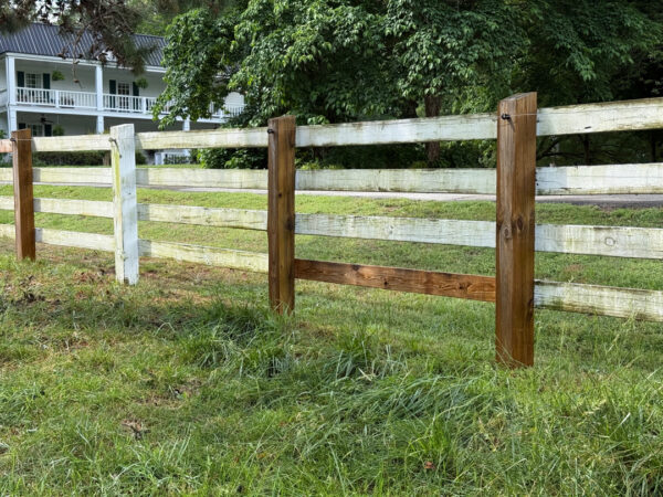 Wooden split-rail fence with brown posts and white rails in a grassy yard, with a white house visible in the background.