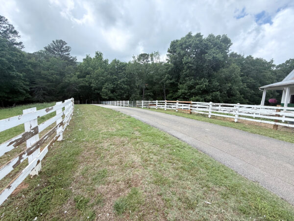 Gravel country road curves between white split-rail fences, with green grass shoulders and a tree-lined background under a cloudy sky.