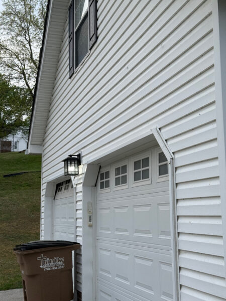 White vinyl-siding garage with two white doors, a black outdoor lantern, and a brown Trinity Disposal trash bin outside the door.